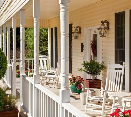 A cozy porch with white rocking chairs, potted plants, and decorative lanterns, surrounded by greenery.