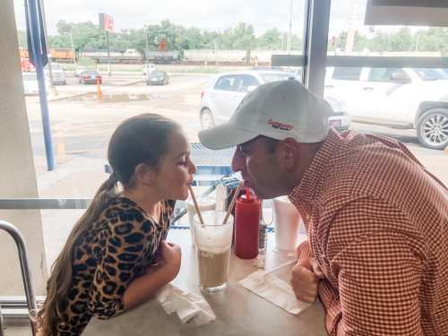 A man and a girl share a milkshake, leaning in with straws touching, smiling at each other in a casual diner setting.