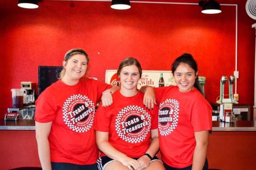 Three young women in red "Treats & Treasures" shirts pose together in a diner with a red wall and soda fountain behind them.