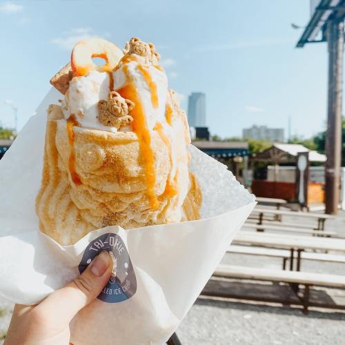 A hand holds a large ice cream cone topped with whipped cream, caramel, and crunchy toppings against a sunny backdrop.