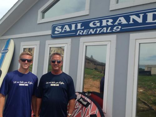 Two men in matching blue shirts stand in front of a rental shop with surfboards and a sign that reads "Sail Grand Rentals."
