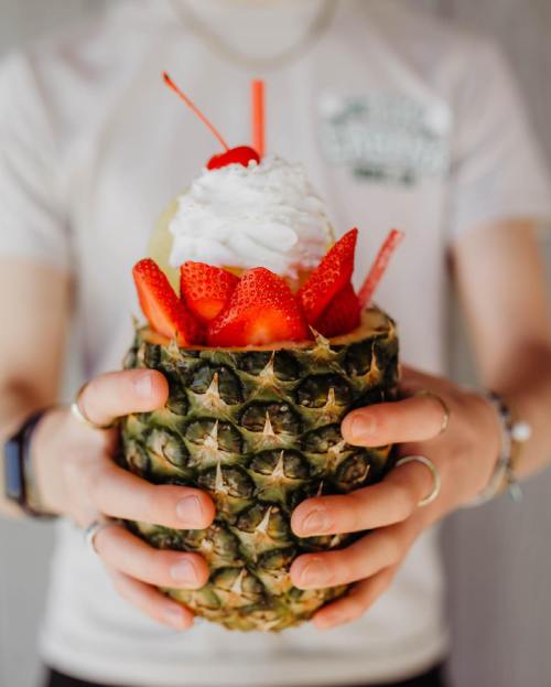 A person holds a pineapple filled with whipped cream, strawberries, and a cherry, with colorful straws.