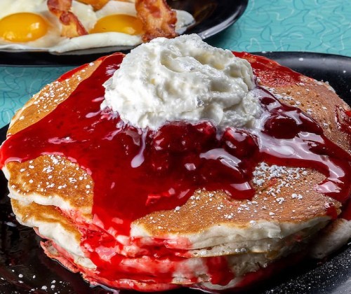 Stack of pancakes topped with whipped cream and strawberry sauce, served on a black plate.