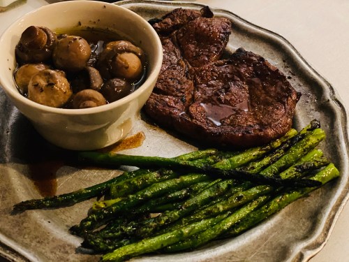 A plate with grilled asparagus, a steak, and a bowl of sautéed mushrooms in olive oil.
