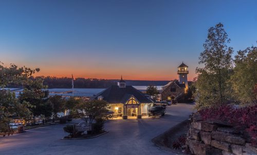 Scenic view of a lakeside marina at sunset, featuring a lighthouse and charming buildings surrounded by trees.