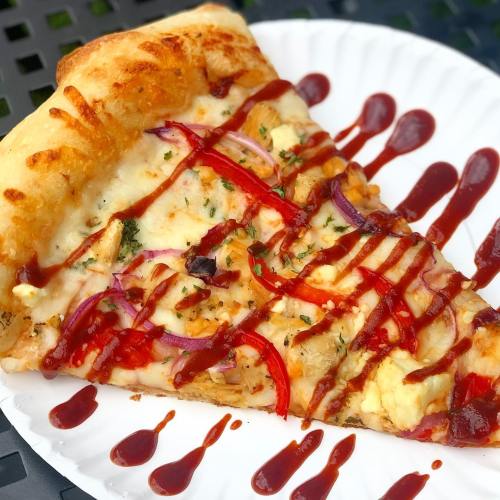 A slice of pizza topped with cheese, red onions, herbs, and drizzled with ketchup on a paper plate.