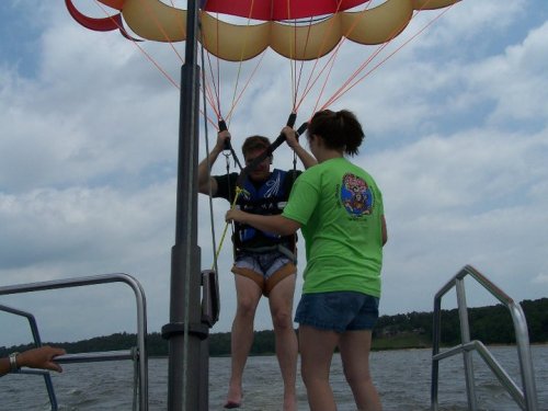 A person in a harness prepares for parasailing while another assists on a boat, with a cloudy sky in the background.