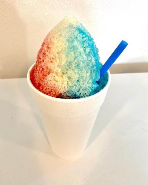 A colorful snow cone in a white cup, featuring red, white, and blue layers, with a blue straw.