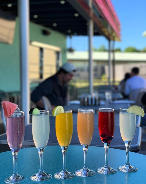A row of colorful cocktails in tall glasses on a table, with a blurred outdoor setting in the background.