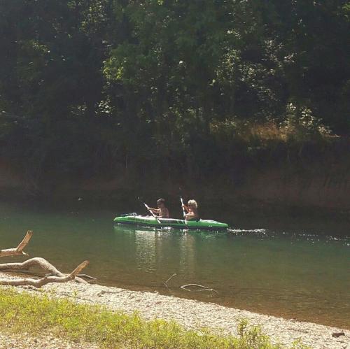 Two people paddle a green kayak on a calm river surrounded by trees and a sandy bank.