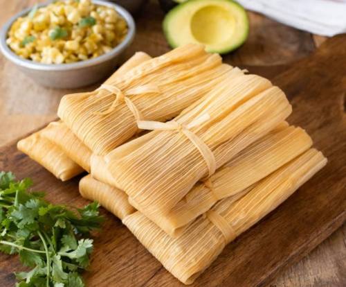 A stack of wrapped tamales on a wooden board, with avocado, cilantro, and a bowl of corn salad in the background.