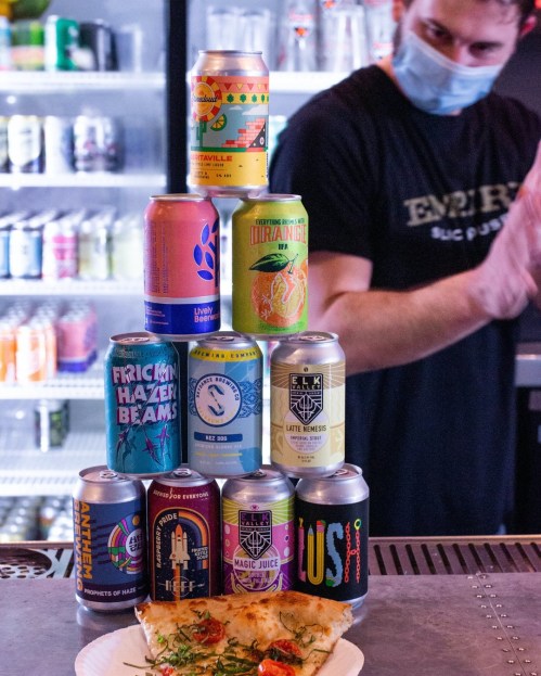 A stack of colorful beer cans next to a slice of pizza, with a masked bartender in the background.