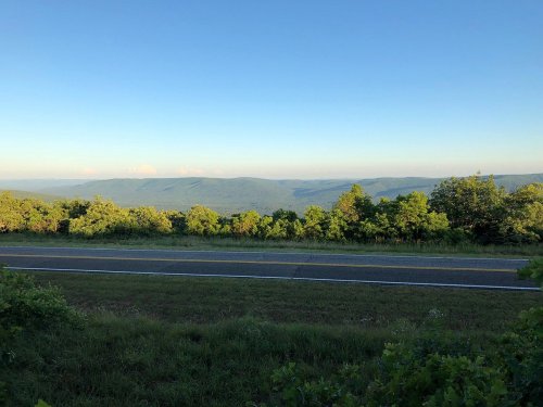 A scenic view of rolling mountains under a clear blue sky, with a road and lush greenery in the foreground.
