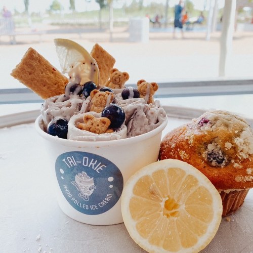 A cup of rolled ice cream topped with blueberries, cookie pieces, and a lemon slice, alongside a muffin and lemon half.