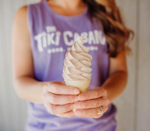 A person holds a soft-serve ice cream cone, wearing a purple tank top with "The Tiki Cabana" printed on it.