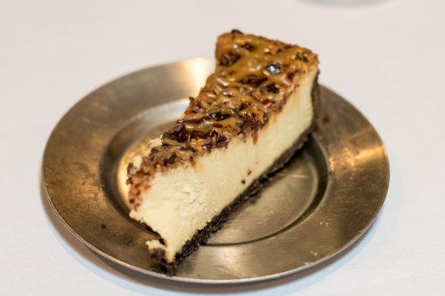 A slice of cheesecake topped with caramel and nuts on a silver plate.