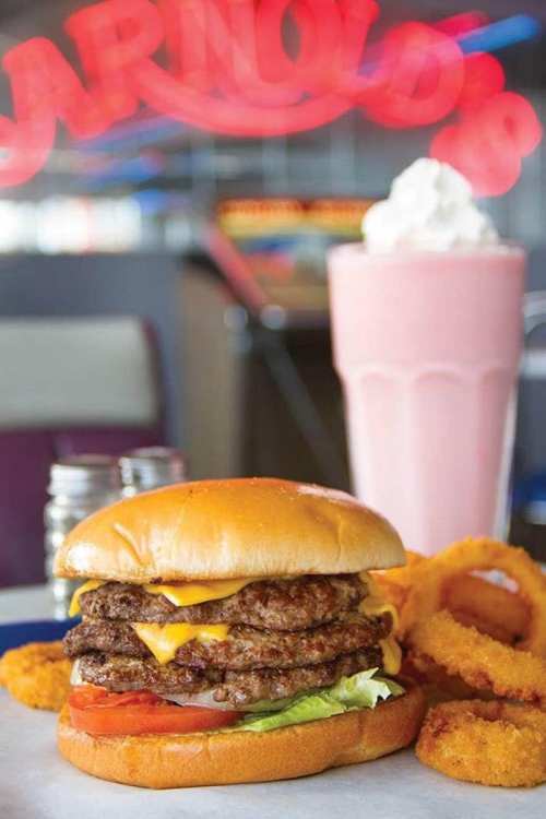 A close-up of a juicy burger with two patties, cheese, lettuce, and tomato, alongside onion rings and a pink milkshake.