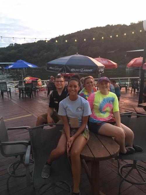 A group of four friends sitting at a table by a lake, with umbrellas and a scenic background at sunset.