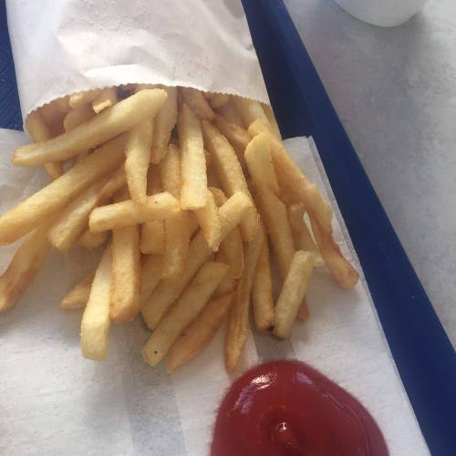 A serving of golden French fries in a paper cone, accompanied by a small dollop of ketchup on a white surface.