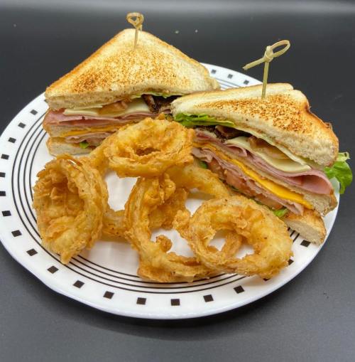 A toasted sandwich with layers of meats and cheese, served with crispy onion rings on a patterned plate.