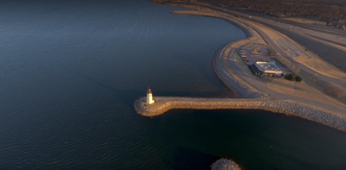 Aerial view of a lighthouse on a rocky pier, surrounded by calm water and a shoreline in the background.
