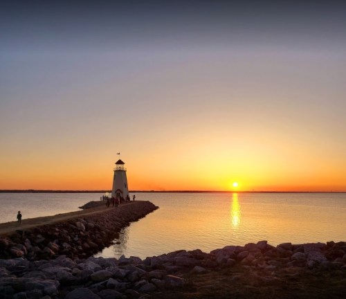 A lighthouse stands on a rocky pier at sunset, with vibrant orange and yellow hues reflecting on the calm water.