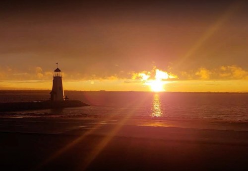 A lighthouse stands by the shore as the sun sets, casting a warm glow over the water and sky.