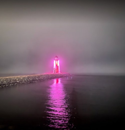 A foggy scene featuring a lighthouse illuminated in pink, reflecting on the calm water.