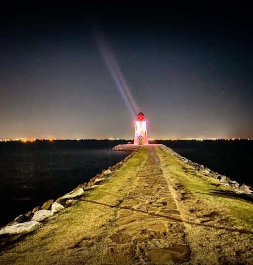 A lighthouse shines brightly at night, standing on a rocky path extending into the dark water, with city lights in the background.