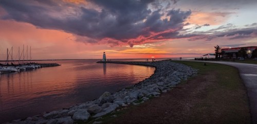 A serene waterfront at sunset, featuring a lighthouse, boats, and colorful clouds reflecting on the water.