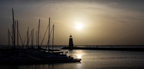Silhouette of a lighthouse at sunset, surrounded by sailboats and calm waters.