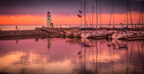 A serene marina at sunset, featuring a lighthouse, sailboats, and colorful reflections on the water.