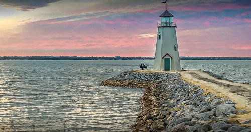 A lighthouse stands on a rocky pier at sunset, with a colorful sky reflecting over the calm water.
