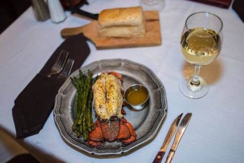 A plate with lobster tail, asparagus, and a small dish of sauce, accompanied by a glass of white wine and a bread loaf.