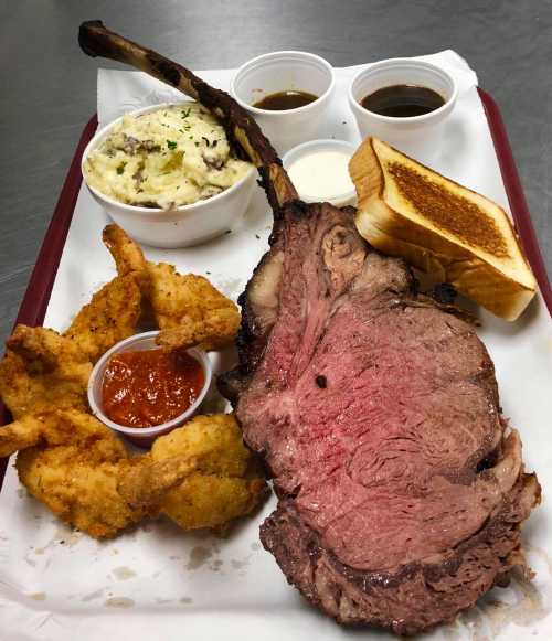 A plate featuring a large prime rib, fried shrimp, mashed potatoes, sauces, and a slice of toasted bread.