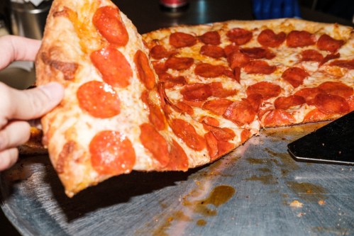 A hand lifts a slice of pepperoni pizza from a large pizza on a metal tray.