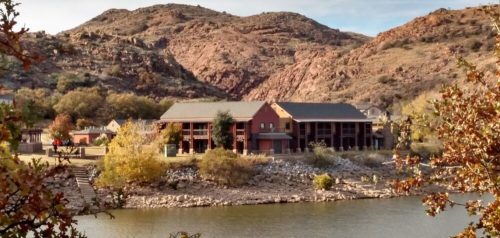 A scenic view of a building by a lake, surrounded by rocky hills and autumn foliage.
