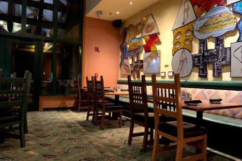 Interior of a restaurant featuring colorful wall art and empty wooden tables with chairs. Soft lighting creates a cozy atmosphere.