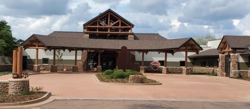 A rustic building with a wooden entrance and stone accents, surrounded by landscaped greenery under a cloudy sky.