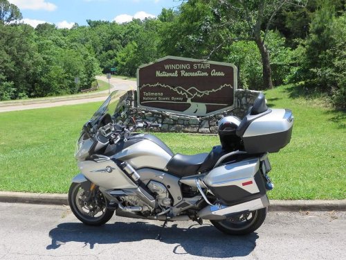 A silver motorcycle parked near a sign for Winding Stair National Recreation Area, surrounded by green trees.