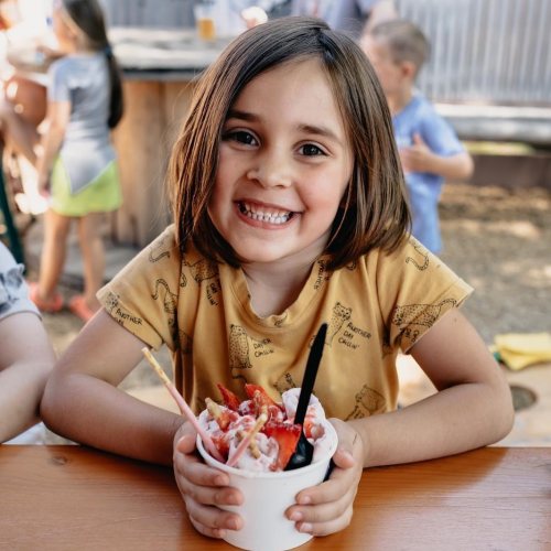 A smiling child with short brown hair holds a cup of colorful ice cream, sitting at a picnic table outdoors.
