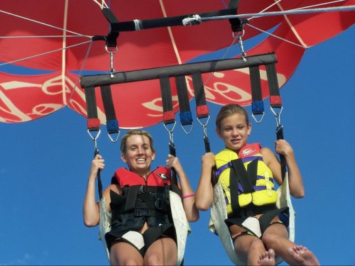 Two girls in life vests smiling while parasailing under a colorful red and white canopy against a blue sky.