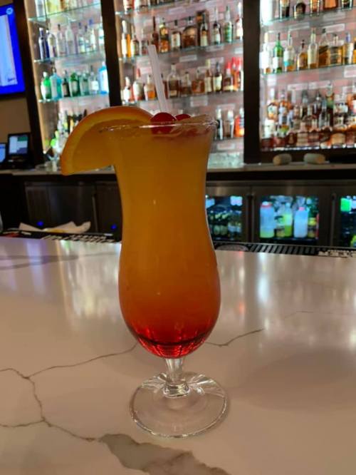 A colorful cocktail in a tall glass, featuring a gradient of orange and red, garnished with a cherry and an orange slice.