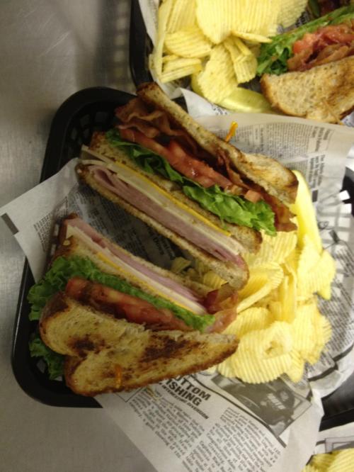 A stacked sandwich with ham, lettuce, and tomato, served with a side of potato chips in a basket lined with paper.