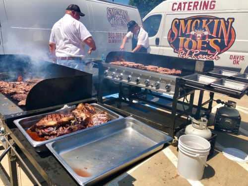 Two chefs grilling meat outdoors at a catering event, with smoke rising and a food truck in the background.