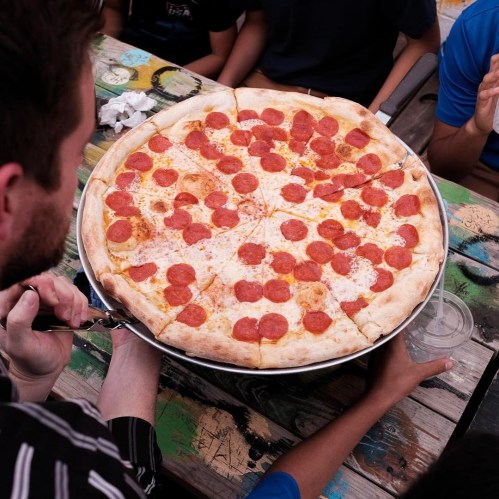 A large pepperoni pizza being served at a table with people in the background.