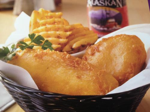 A basket of golden fried fish fillets with crinkle-cut fries and a drink in the background.