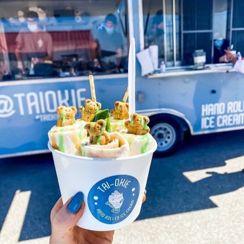 A hand holding a cup of rolled ice cream topped with colorful toppings, in front of a food truck.