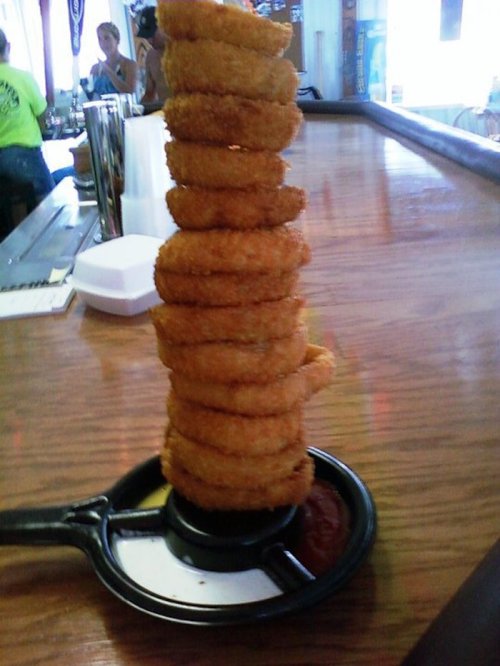 A tall stack of golden onion rings on a black plate with a side of ketchup, set on a wooden bar counter.