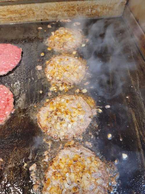 Sizzling burgers with onions cooking on a griddle, steam rising in a kitchen setting.